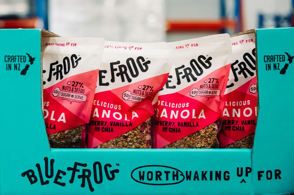 Packets of Blue Frog raspberry, vanilla and chia granola displayed in a teal box.