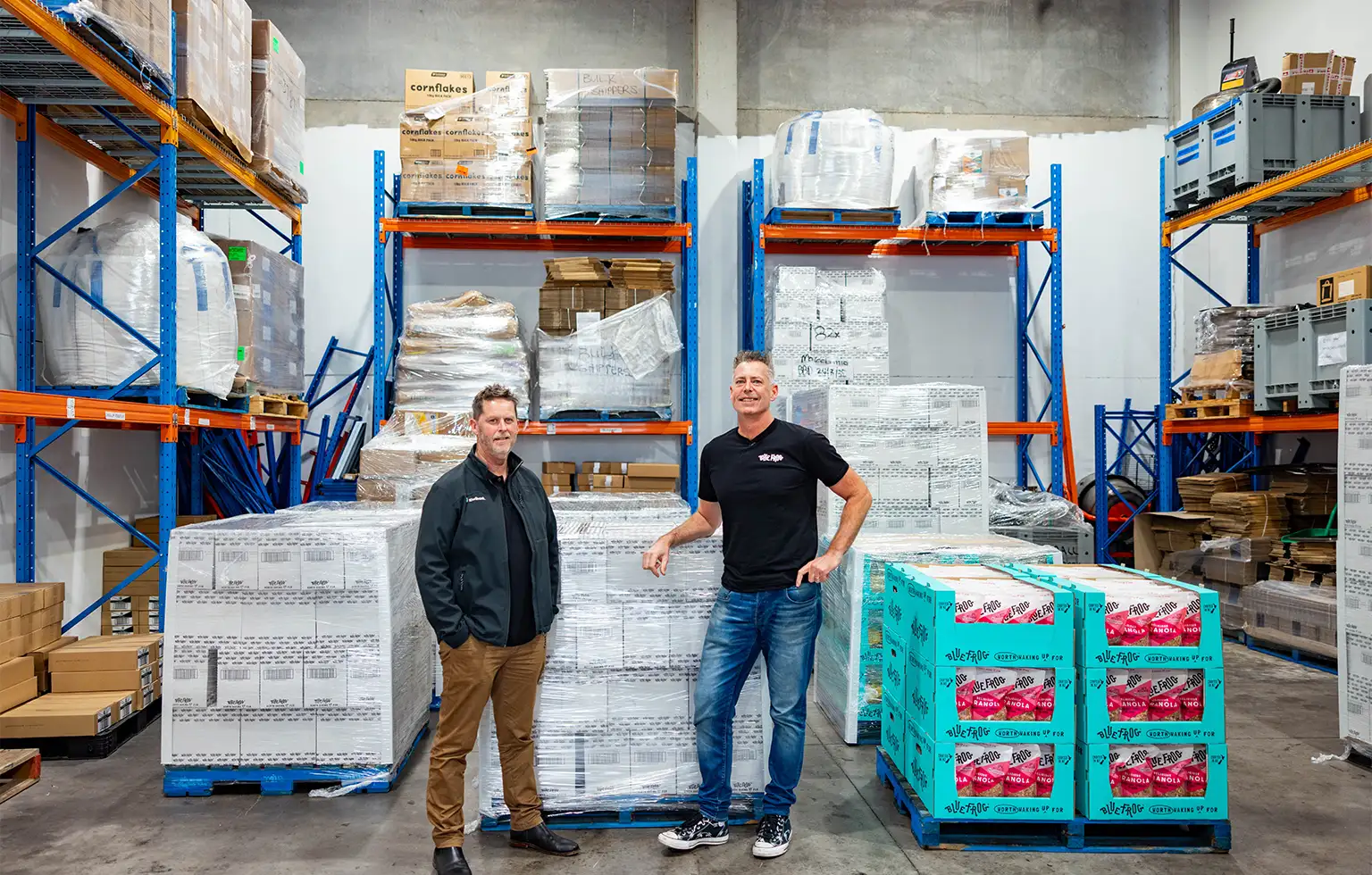 Two men standing in a warehouse surrounded by stacked pallets of packaged food products.