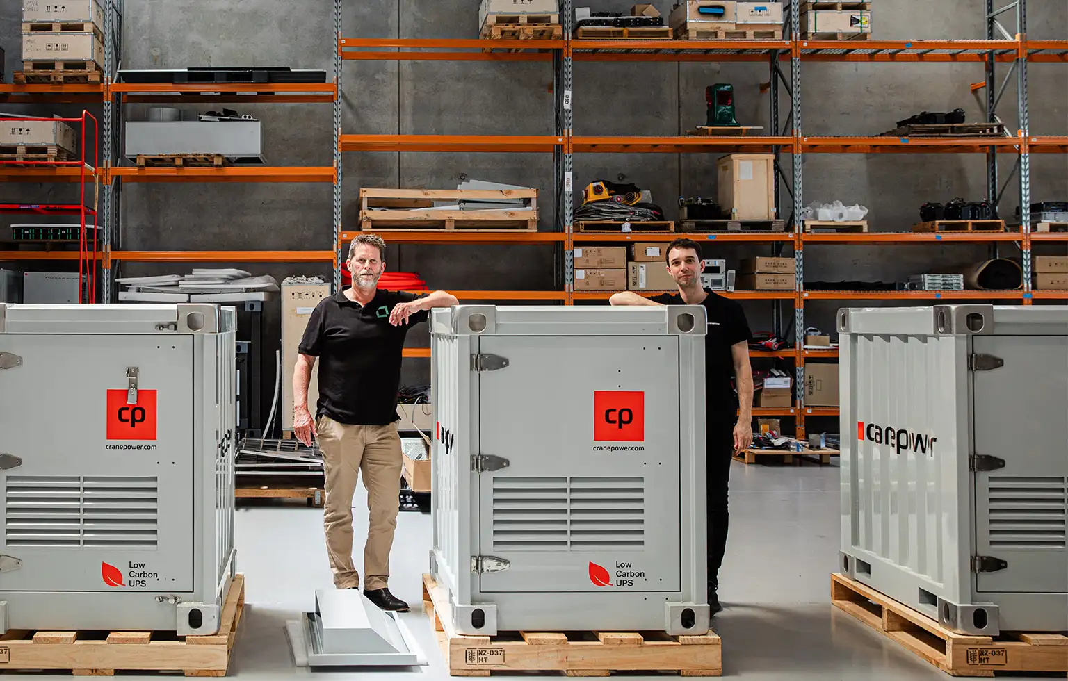 Two people standing beside large grey Cranepower units inside a warehouse.