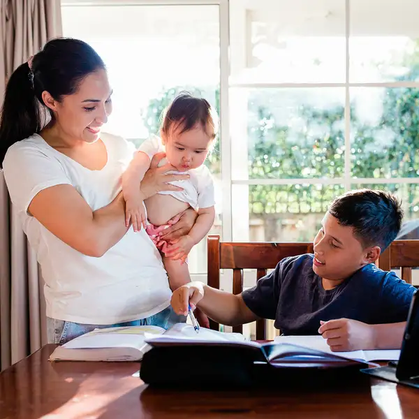 Parent holding baby while child doing homework dining table
