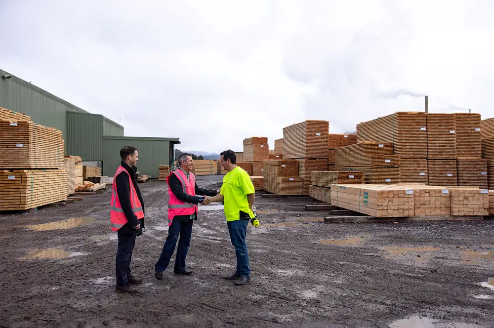 Three people standing in a timber yard, with two shaking hands among stacks of lumber.