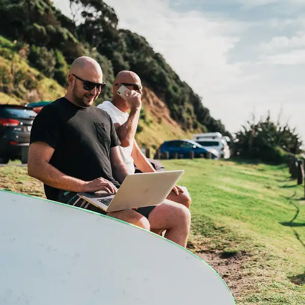 Person uses laptop beside surfboard on grassy coastline sunny setting