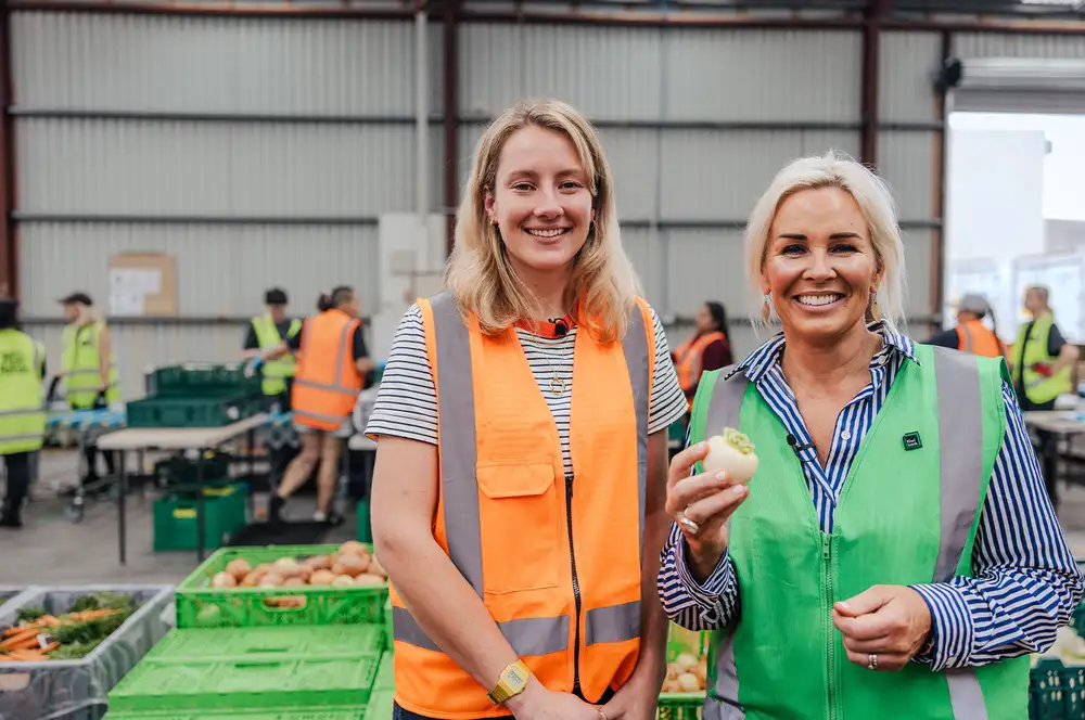 Two people in high‑visibility vests standing in a produce warehouse, one holding a vegetable.