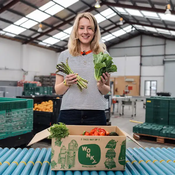 A person holding fresh produce beside an open Wonky Box filled with vegetables in a warehouse.