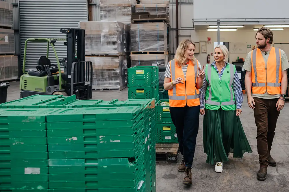 Three people in high‑visibility vests walking through a produce warehouse beside stacks of green crates.